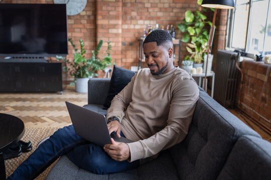 Happy young adult male sitting on sofa in a loft apartment working on a laptop