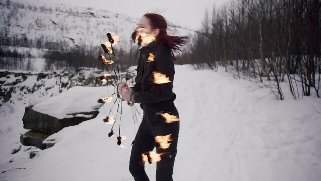 A young redhead caucasian girl in a black shirt and black jeans masterly spins staff fire fans and than close her eyes in the winter forest with snow, hills and trees. Perfomance in a fire show