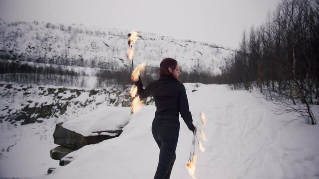 A young redhead caucasian girl in a black shirt and black jeans masterly spins staff fire fans in the winter forest with snow, hills and trees. Perfomance with staff in a fire show