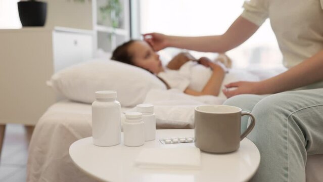 Focus on bedside table with cup of hot beverage, paper tissues and medical pills standing near white bed. Blurred background of caring mother measuring daughter's fever with digital thermometer.