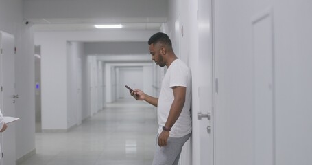 African American man stands in hospital corridor, records voice message and chats by mobile phone. Recovering patient during treatment in modern clinic. Medical staff walk in background. Slow motion.