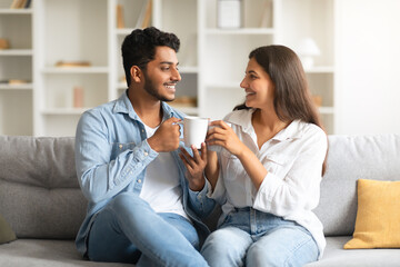 Happy indian couple talking and drinking coffee, sitting on sofa