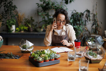 Asian florist talking on smartphone near notebook, succulents and terrariums in floral store,...