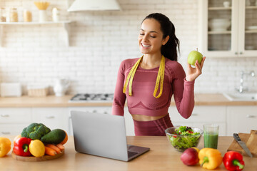 Smiling fitness nutritionist lady using laptop holding apple in kitchen