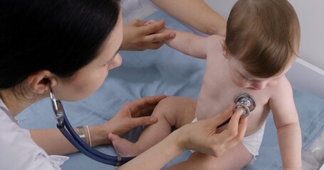 Mother holds hand of cute baby with pacifier sitting on changing table. Doctor uses stethoscope to...
