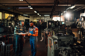 A wide shot of two factory workers in a hangar with various parts and machines.