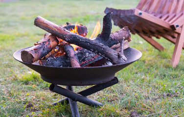 Metal fire bowl with burning wood in the yard and garden rack lounge chair. Safe decorative fire...