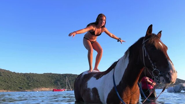 Young red-haired girl in bikini enjoys standing on horse bathing in sea water in summer season