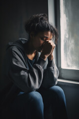 African American depressed sad teenage girl in grey hoodie sitting next to the window crying