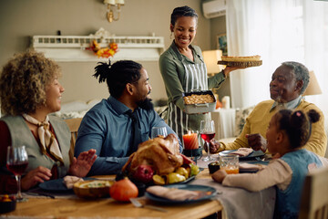 Fototapeta na wymiar Happy black woman serving Thanksgiving dessert to her family at dining table.