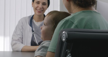 Obraz premium Female pediatrician consults patient with cute little child on appointment in hospital. Woman sits on chair at physician checkup. Friendly doctor works in modern medical center. Close up. Back view.