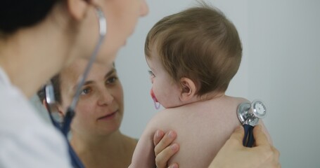 Happy mother holds on hands her handsome baby with pacifier in mouth. Pediatrician uses stethoscope to listen to heartbeat and lungs of cute little child. Physician does checkup in hospital. Close up.