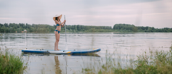 Fototapeta na wymiar A young woman with an open swimsuit swims on a SUP board on a picturesque lake. Evening tour. A beautiful, slender girl is engaged in sap surfing on the calm water of a picturesque pond.