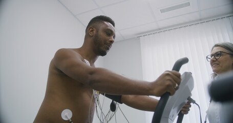 Female cardiologist, doctor puts holter sensors on patient with cardiovascular disease. African American man during functional rehabilitation in cardiology room. Electrocardiography in modern hospital