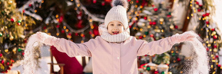 Little girl in winter clothes on standing cheerful on a decorated Christmas street in winter concept of Christmas or New Year greetings. Banner with copy space.