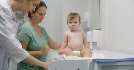 Mother holds baby on hands. Medic puts scale on changing table. Female doctor weighs kid in modern...