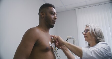 Female cardiologist, doctor puts holter sensors on patient with cardiovascular disease. African American man during functional rehabilitation in cardiology room. Electrocardiography in modern hospital