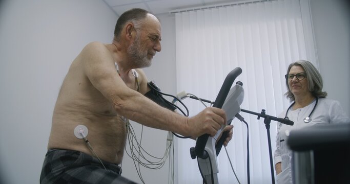 Elderly man with holter sensors pedals orbitrek in cardiology room. Female cardiologist does diagnostic of recovering patient with cardiovascular disease. Functional rehabilitation in modern hospital.