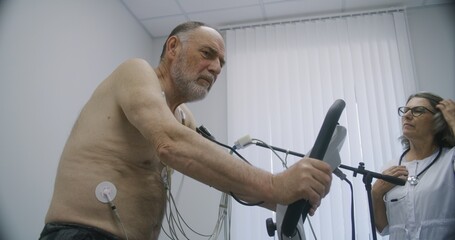 Elderly man with holter sensors pedals orbitrek in cardiology room. Female cardiologist does...