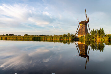 Typical Dutch windmill in the Kinderdijk area, the Netherlands with a near perfect reflection of the windmill, reed beds and clouds in the tranquil water during last hours of the day - obrazy, fototapety, plakaty