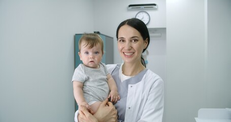 Female pediatrician stands in middle of bright hospital room and looks at camera. Adult doctors holds baby. Little boy looks at camera and holds hand of medic. Medical staff at work in modern clinic.
