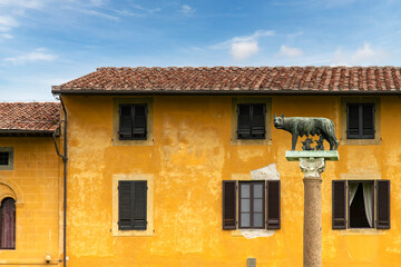 The Capitoline Wolf (Lupa Capitolina) bronze sculpture on Piazza del Duomo in Pisa, Italy against the backdrop of yellow plastered façade of typical Italian houses