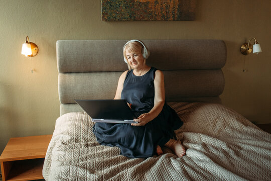 An Elderly Woman Wearing Headphones Communicates Online Or Watches A Video On A Laptop In The Bedroom At Home