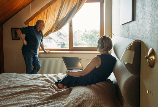 An elderly woman communicates online, watches videos or works on a laptop. An elderly man is standing at the window.
