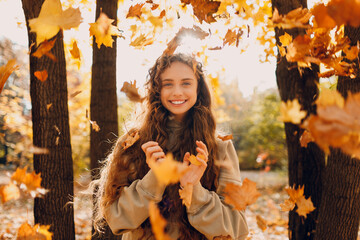 Smiling positive young woman throws up the yellow autumn leaves in the forest at sunset