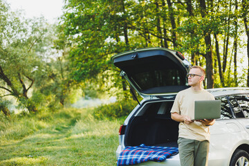 Cheerful traveler, young man sitting in open trunk of car, working with laptop, outdoors, concept of business, travel, vacation