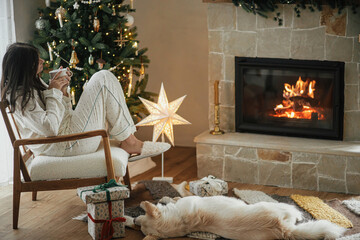 Cozy christmas morning. Beautiful woman in stylish pajamas relaxing with warm tea and cute dog at...