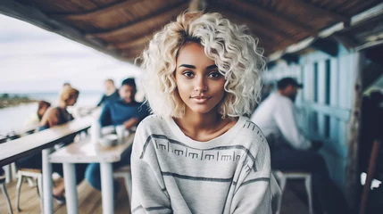 Foto auf Acrylglas young adult woman, tanned skin tone, 20s 30s, dyed blonde curly hair, wearing thin cozy casual sweater, outside on a pier or harbor with wooden jetty or restaurant, waiting © wetzkaz