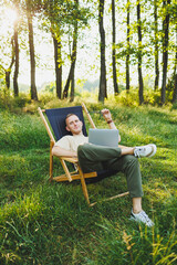 Obraz premium A man works on a laptop while sitting on a bench in a city park. A young guy reads the news, writes a message on his laptop. Recreation in nature, remote work on vacation