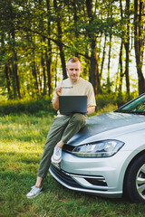 A cheerful man in glasses sits on the hood of his car in nature and works on a laptop. Freelance work, remote work during vacation