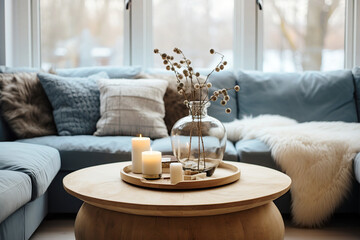 Glass vase with twig on round wooden coffee table near cozy blue sofa with fur blanket and knitted pillows. Scandinavian hygge home interior design of modern living room. - obrazy, fototapety, plakaty