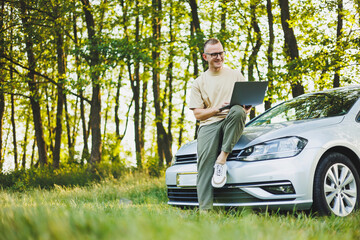 A successful man in glasses works on a laptop while leaning on the hood of his car. Remote work in nature. Work on a laptop online. Working on a computer while traveling.