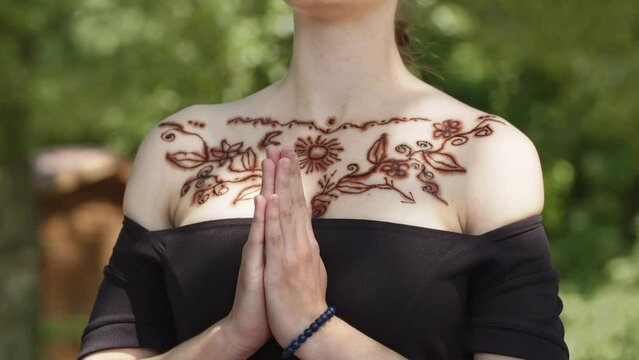 A girl in black dress with henna tattoo on chest within an Indian pattern. A young woman at oriental holiday or festival meditates, prays and folds her palms near her chest. Gesture of peace and