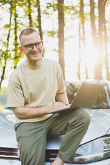 Fototapeta premium A serious man in glasses is working on a laptop on the hood of his car. Remote work in nature. Work on a laptop online. Working on a computer while traveling.