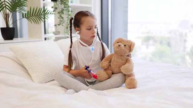 Little caucasian girl listening heartbeat of teddy bear with toy stethoscope in cozy bed against background of panoramic window with city view. Concept of childhood, game and treatment.