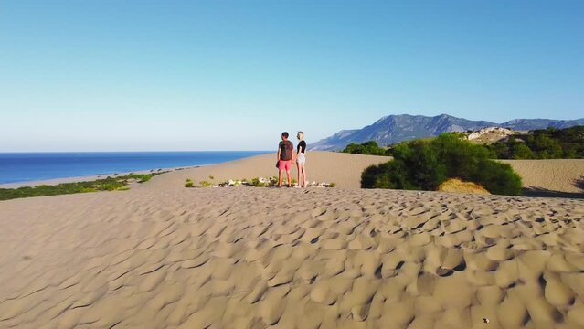 Drone flies around circumference of pair of happy travelers walking along sand dunes on beautiful Patara beach on Mediterranean coast. Wanderlust, social media content. Turkey Mountains and seas. Heat