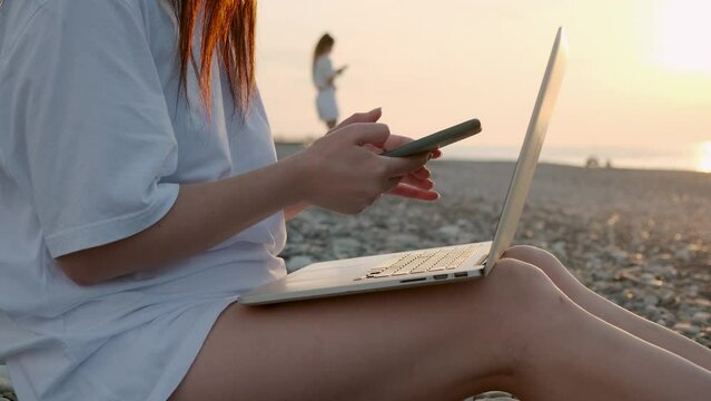Close up of unrecognizable woman working with laptop and mobile phone on the beach by the sea during sunset. Female freelancer work online using smartphone and computer from a distance