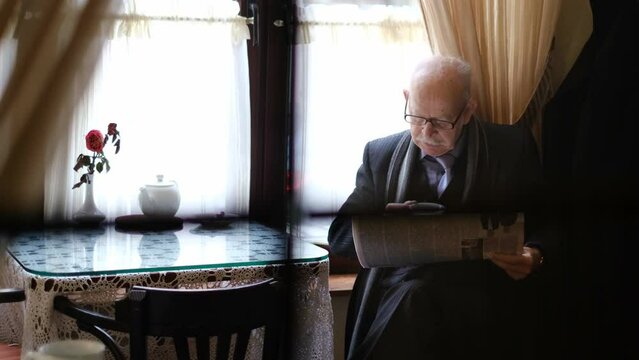 Elderly pensioner in glasses reading a daily newspaper, sitting in a coffee shop and drinking coffee.