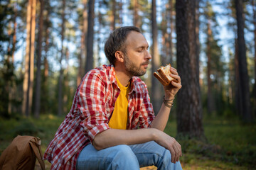 Fototapeta premium Carefree man eating sandwich snack during hike walk travel camping vacation, feels calm relax in nature landscape. Happy hiking male resting taking break with healthy food in forest. Autumn nature.