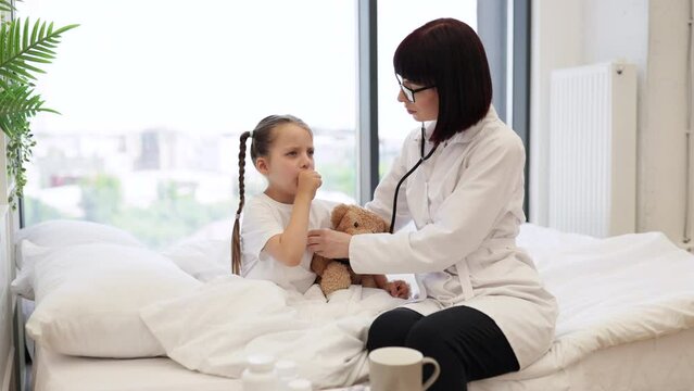 Experienced nurse sitting on bed and using stethoscope while small lady covering mouth with hand and coughing in bedroom. Concept of home treatment and friendly relations between kid and doctor.