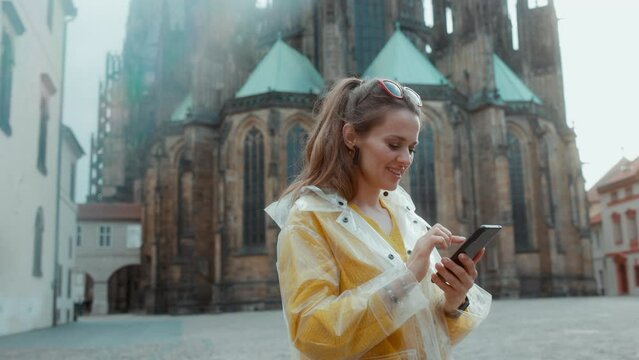 surprised stylish woman in yellow blouse and raincoat in Prague Czech Republic enjoying promenade and using smartphone.