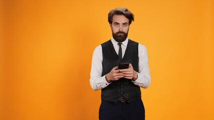 Stylish waiter uses smartphone to text before serving customers at table, browsing online pages against yellow background. Professional valet working at fine dining restaurant.