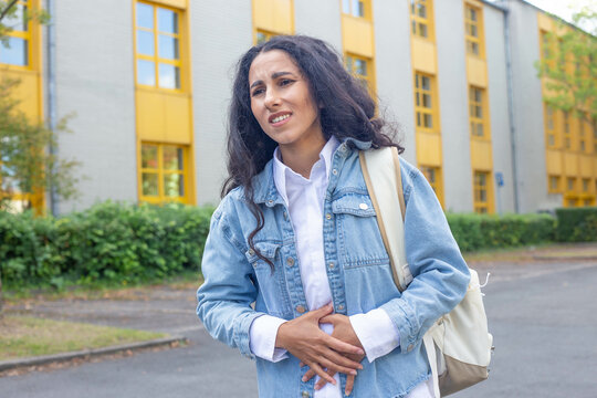 A young Spanish woman on the street near the building of a school or hospital experiences a sharp pain in the abdomen, holds with both hands the lower abdomen of the antics, feels a sharp spasm.