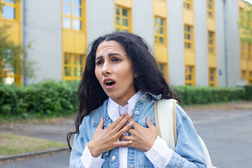 Young Eastern girl suddenly felt a panic attack, shock, suffocation, holding her chest with her hands and breathing heavily. Against the background of a hospital or a student on a university campus. - obrazy, fototapety, plakaty