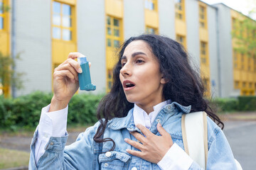 Young Eastern girl has an asthma attack or panic and holds an inhaler in her hand while leaving a school or university building. Inhale vapor from an inhaler - first aid on the street. - obrazy, fototapety, plakaty