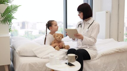 Competent female pediatrician prescribing medicine on modern tablet for sick child in spacious bedroom. Smiling girl holding teddy bear and preparing for intensive treatment at home. - Powered by Adobe